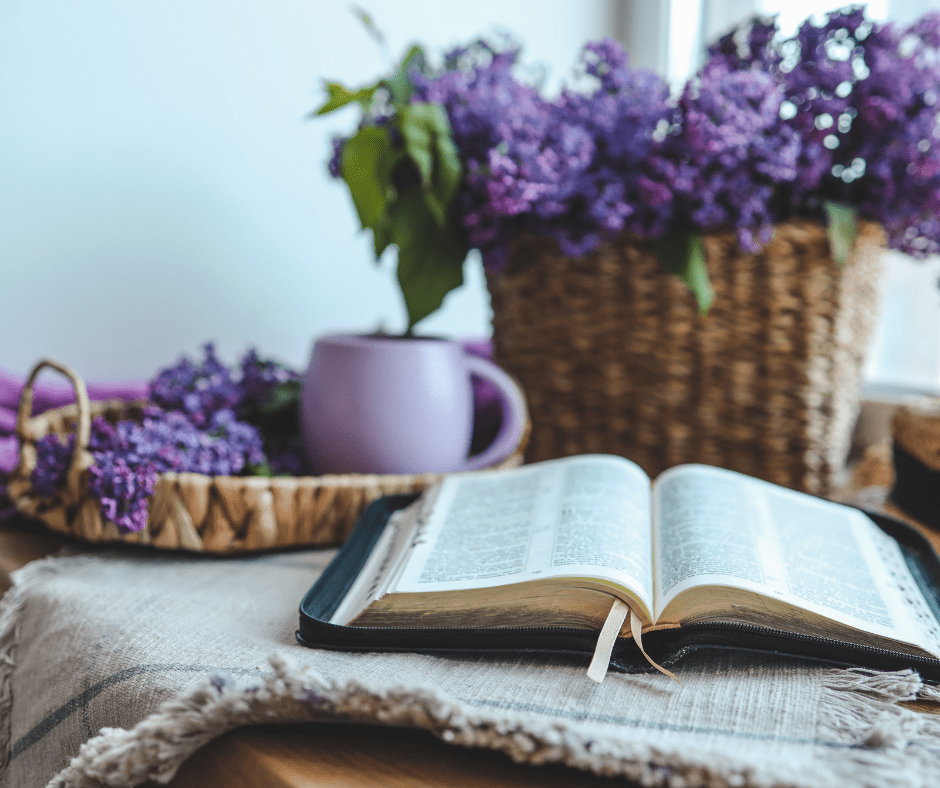 A basket full of purple hydrangea flowers stands behind a bible on a table with a cozt mug.